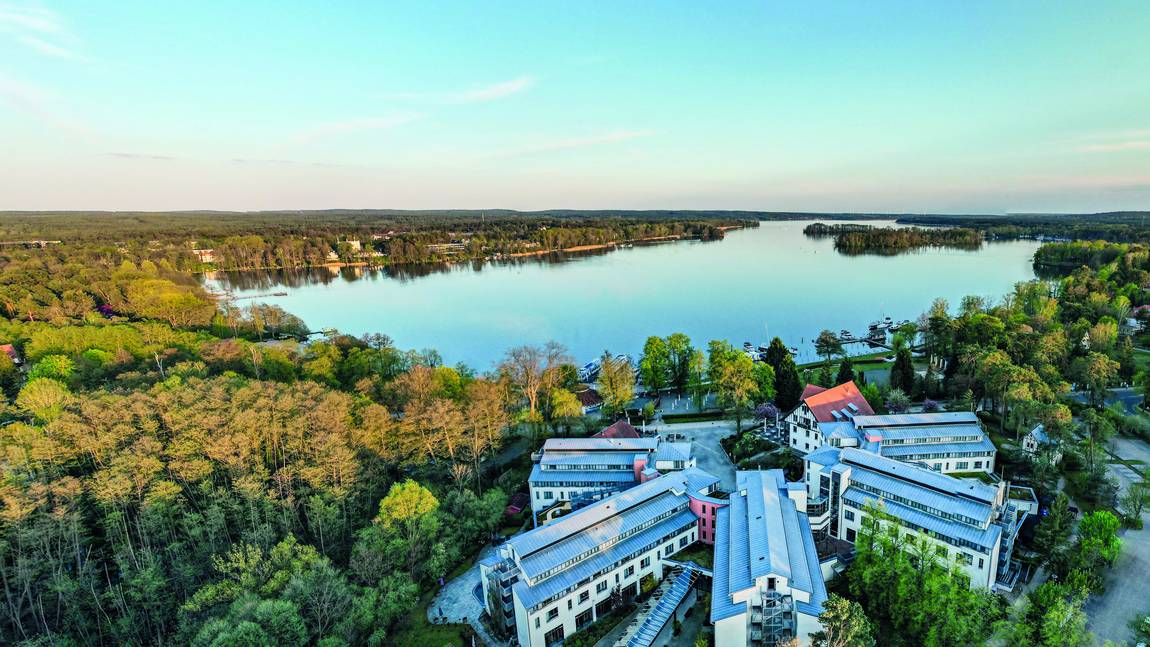 ©Hotel Esplanade Resort & Spa Bad Saarow Luftaufnahme einer Seelandschaft mit ruhigem Wasser, umgeben von üppig grünen Bäumen und Wald. Im Vordergrund sind mehrere moderne Gebäude mit weißen Fassaden in Ufernähe unter einem klaren blauen Himmel zu sehen.