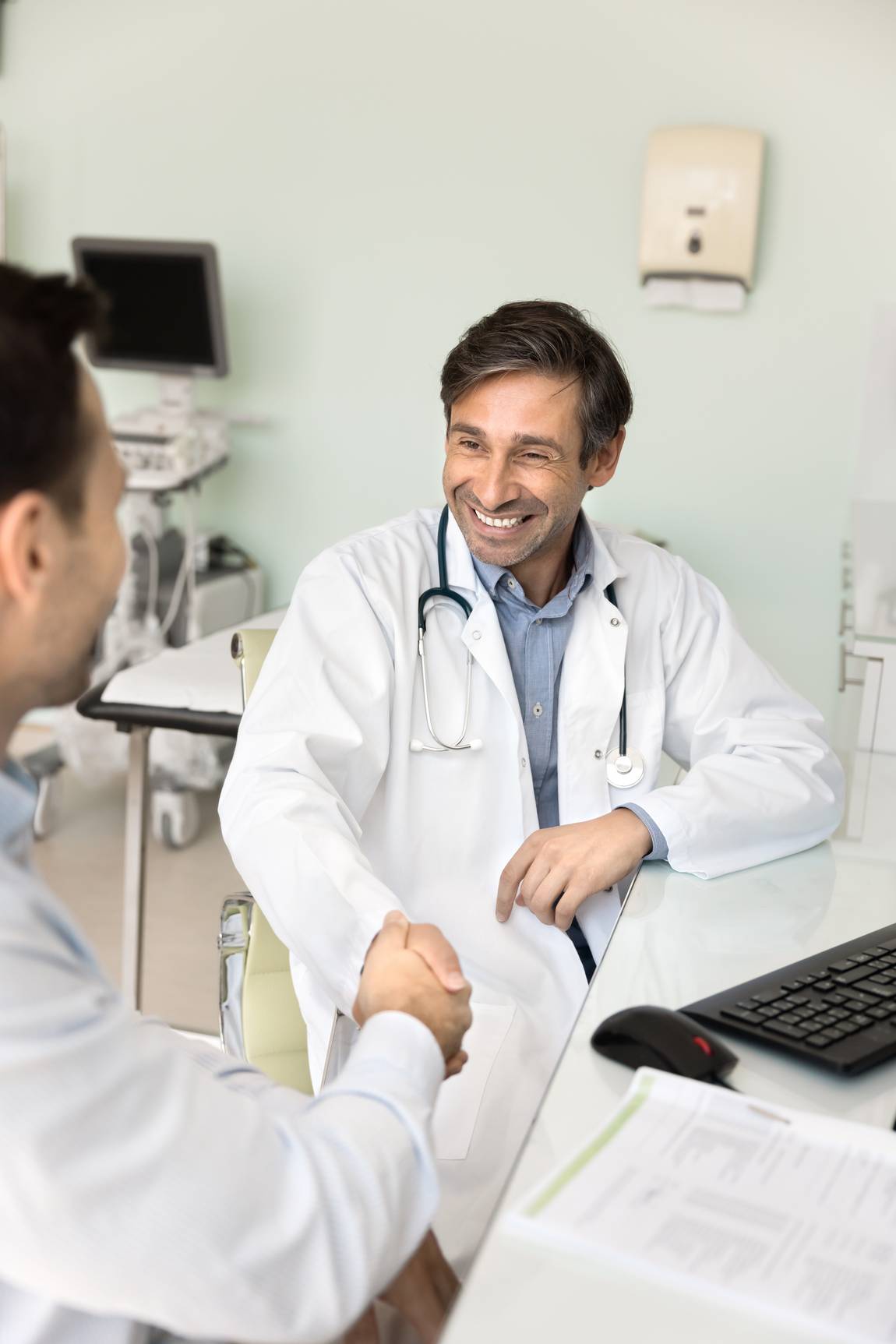 ©istock.com/fizkes Ein lächelnder Arzt mit weißem Kittel und Stethoskop sitzt an einem Schreibtisch und schüttelt einem Patienten in einem medizinischen Büro die Hand. Ein Computer, Papierkram und medizinische Geräte sind im Hintergrund zu sehen.