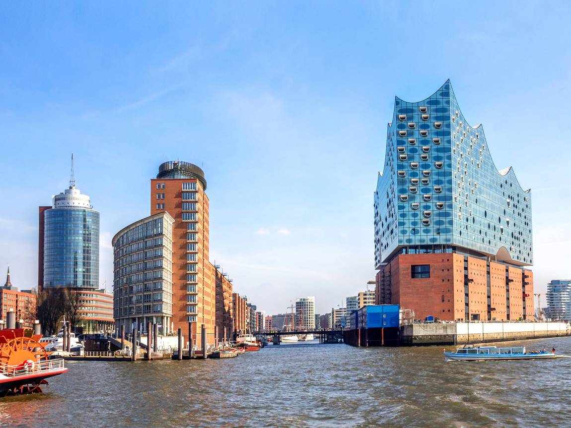 ©pure-life-picturesstock/adobe.stock.com Eine Stadtlandschaft am Wasser mit modernen und historischen Gebäuden, darunter die gläserne Elbphilharmonie in Hamburg, Deutschland, unter einem klaren blauen Himmel, mit Booten auf dem Fluss im Vordergrund.