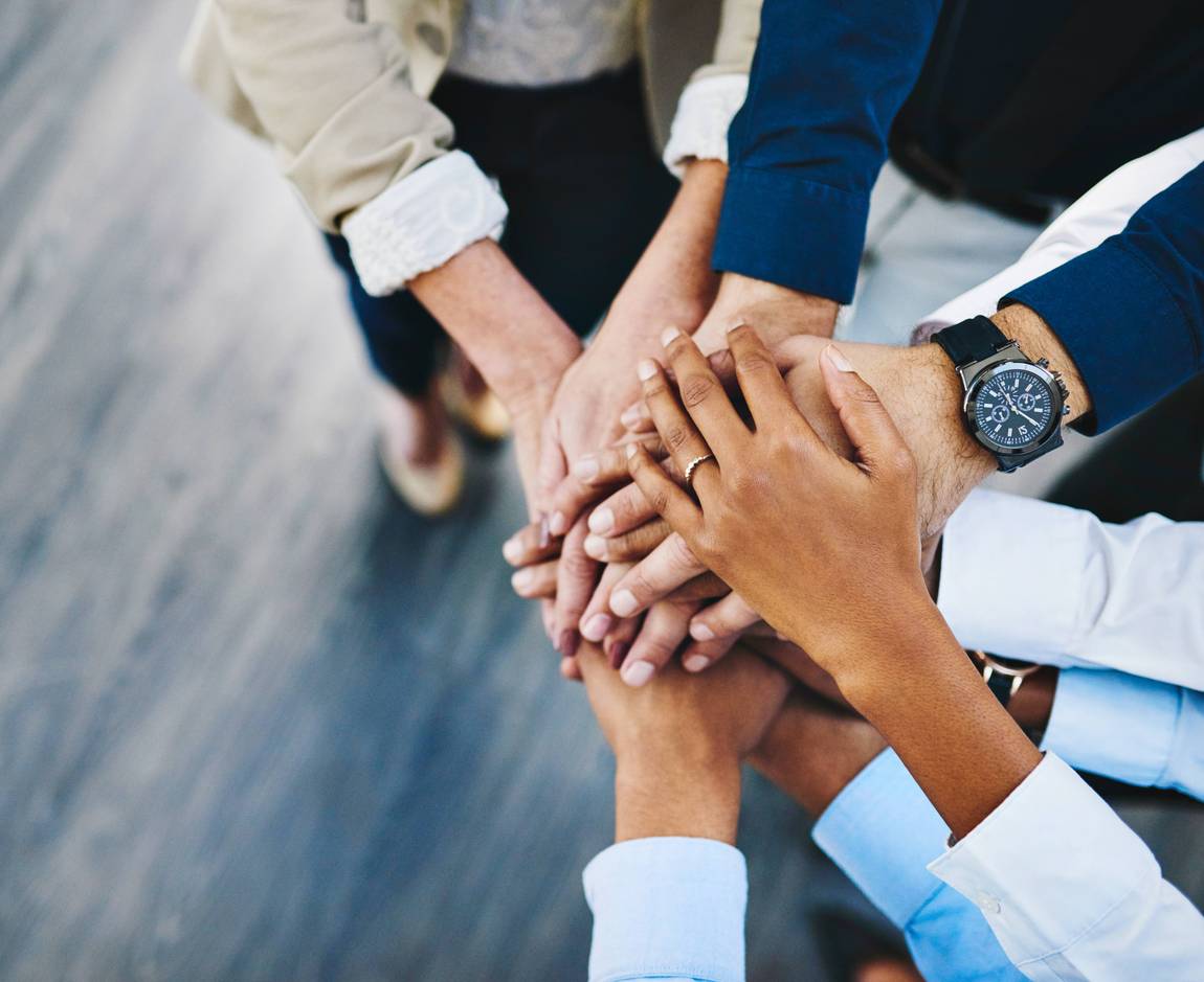 ©PeopleImages.com - Yuri A/Shutterstock.com Eine Gruppe von Menschen, die in einem Kreis stehen, legen ihre Hände in der Mitte zusammen, um Teamwork und Einheit zu symbolisieren. Der Boden unter ihnen ist grau und aus Holz.