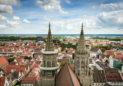 ©istock.com/frahaus Ansicht einer Stadt mit zwei hohen, verschnörkelten Kirchtürmen im Vordergrund, umgeben von Gebäuden mit roten Dächern, Bäumen und einem wolkenverhangenen Himmel über dem Kopf.