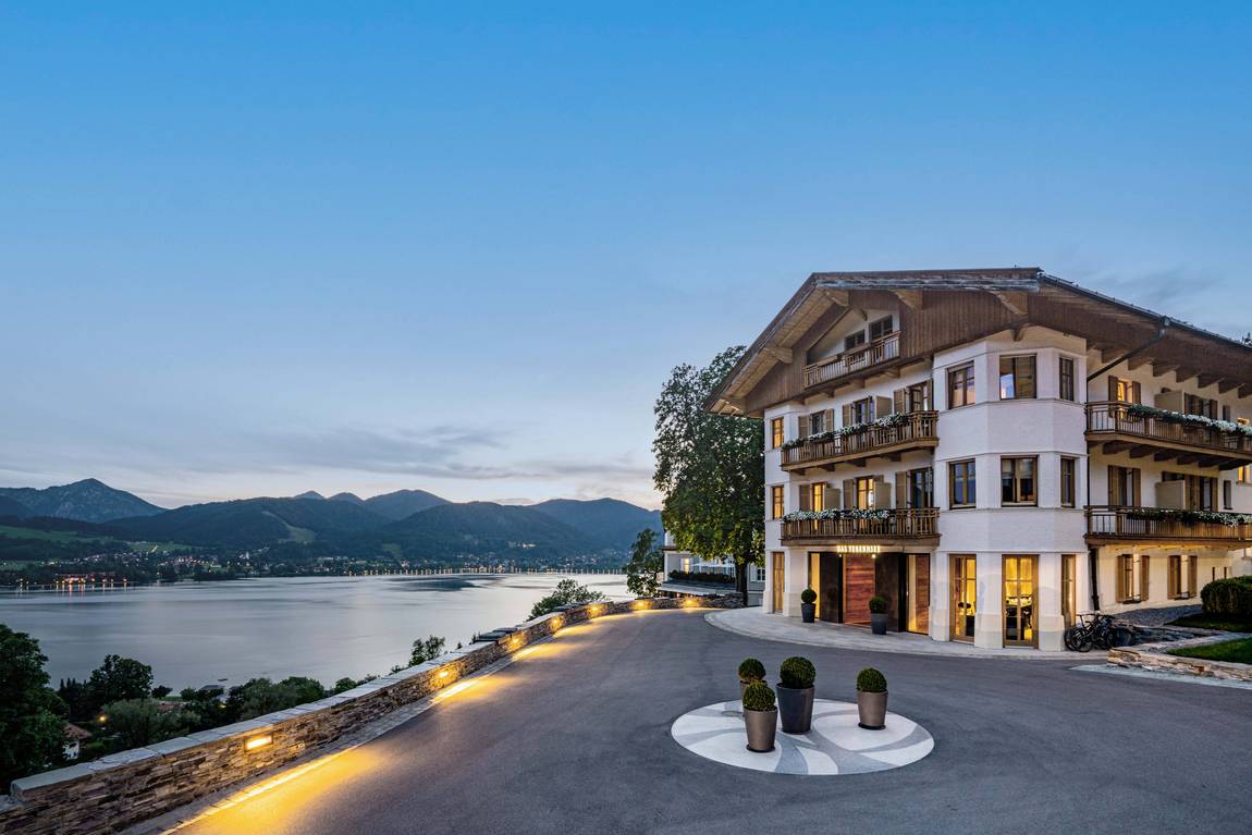 © DAS TEGERNSEE Ein großes, elegantes Hotel mit Balkonen liegt bei Sonnenuntergang auf einem Hügel mit Blick auf einen ruhigen See und ferne Berge, mit einer kreisförmigen Auffahrt und Topfpflanzen im Vordergrund.