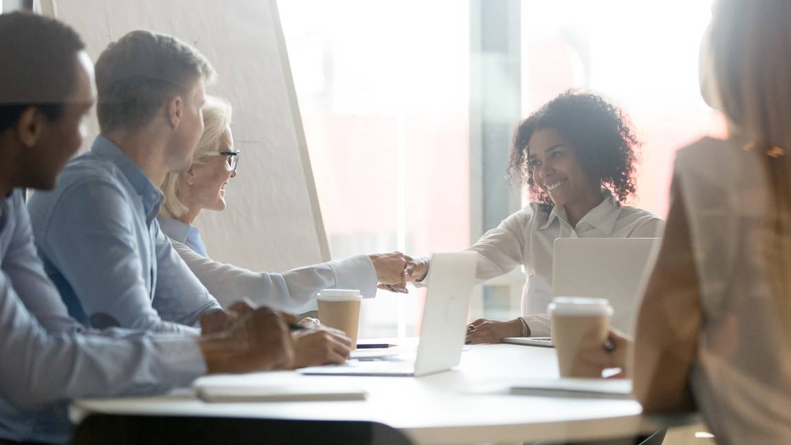 ©istock.com/fizkes Fünf Personen sitzen um einen Tisch in einem Besprechungszimmer. Zwei Frauen in der Mitte lächeln und stoßen sich mit der Faust an. Laptops, Papiere und Kaffeetassen stehen auf dem Tisch, und im Hintergrund ist ein großes Fenster zu sehen.
