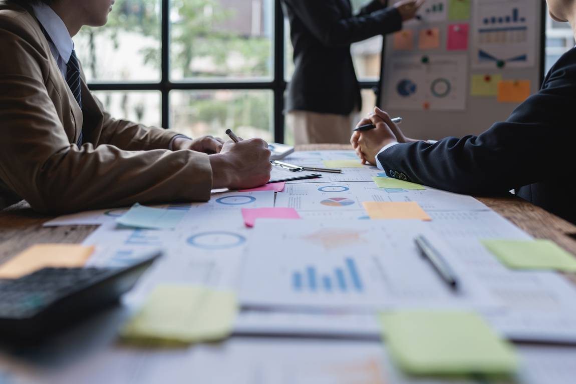 ©istock.com/Worawee Meepian Drei Personen in Geschäftskleidung befinden sich in einem Besprechungsraum mit Diagrammen, Schaubildern und Haftnotizen auf einem Tisch. Eine Person präsentiert an einer Tafel, während zwei andere sitzen und Notizen machen. Helles Fenster im Hintergrund.