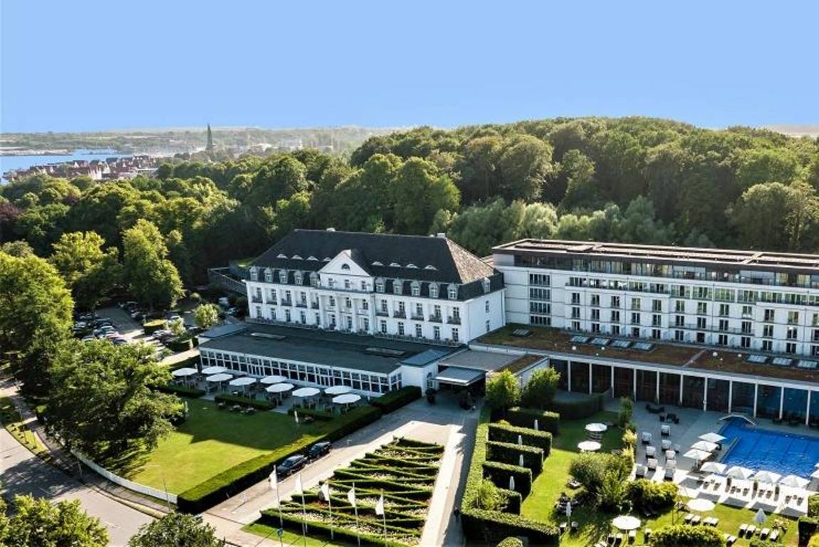 ©A-ROSA Travemünde Luftaufnahme eines großen, eleganten Hotels, umgeben von üppigen grünen Bäumen, mit Gärten, Sitzgelegenheiten im Freien und einem blauen Swimmingpool; im Hintergrund sind eine Stadt und Wasser unter einem klaren blauen Himmel zu sehen.