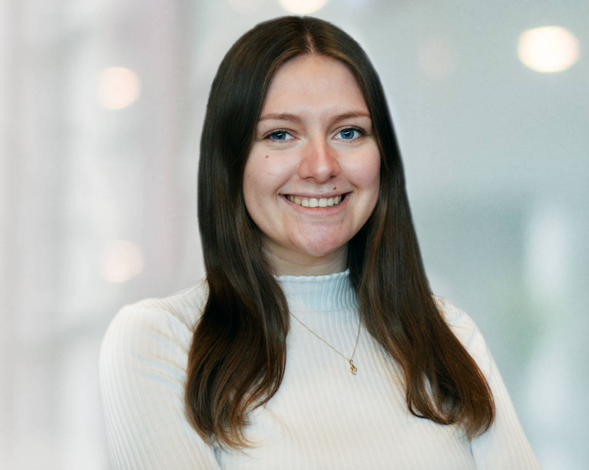 Eine Frau mit langen braunen Haaren, die einen weißen Rollkragenpullover und eine Halskette trägt, lächelt, während sie vor einem verschwommenen, hellen Hintergrund steht.